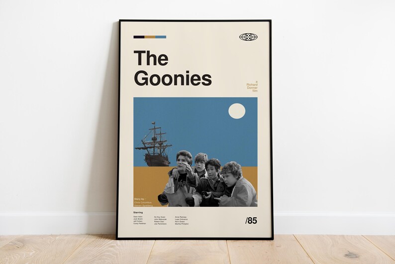 May include: A vintage-style poster for the film "The Goonies", featuring a group of kids looking out at a ship sailing on a blue sea with a white moon in the sky. The poster is in a black frame and has the text "The Goonies" in bold black letters.