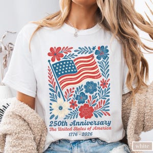 Puede incluir: Camiseta blanca con un dise&ntilde;o patri&oacute;tico. El gr&aacute;fico incluye una bandera estadounidense, rodeada de elementos florales rojos, azules y crema. El texto dice "250th Anniversary The United States of America 1776-2026."