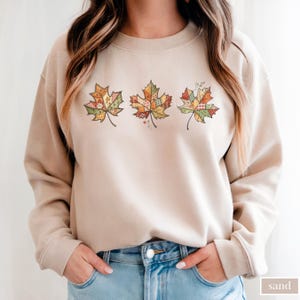 May include: A sand-colored sweatshirt featuring three appliqued maple leaves. Each leaf is a patchwork of autumn-toned fabrics, including orange, green, and yellow. The leaves are arranged horizontally across the chest. The word "sand" is printed in the bottom right corner.