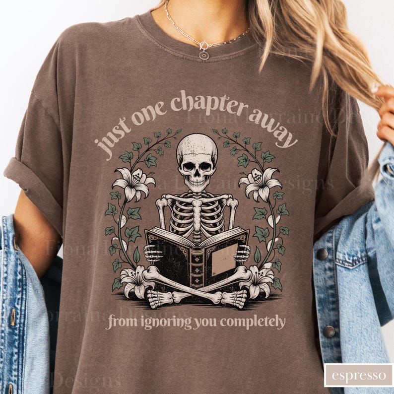 May include: Espresso-colored t-shirt featuring a skeleton reading a book, surrounded by floral elements. The text reads "just one chapter away from ignoring you completely." The design is printed on a soft, comfortable tee.