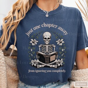 May include: A denim blue t-shirt features a skeleton reading a book, surrounded by floral elements. The text reads "just one chapter away from ignoring you completely." The design is centered on the shirt.