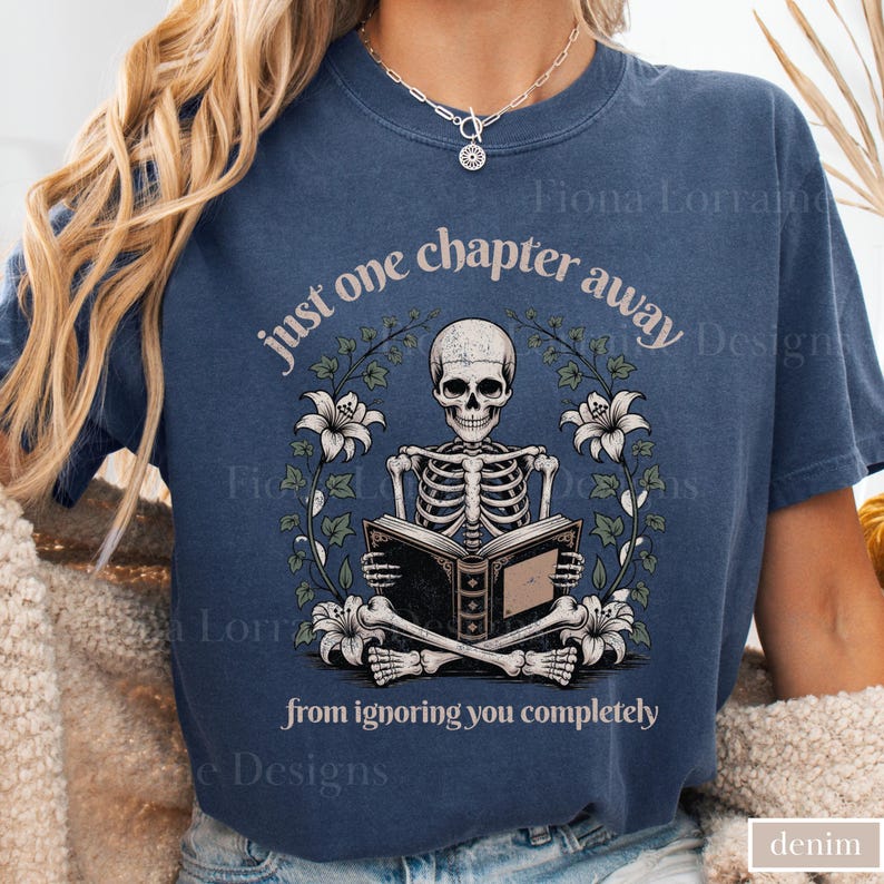 May include: A denim blue t-shirt features a skeleton reading a book, surrounded by floral elements. The text reads "just one chapter away from ignoring you completely." The design is centered on the shirt.