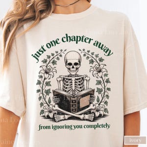 May include: Ivory-colored t-shirt featuring a skeleton reading a book, surrounded by floral and vine accents. The text reads "just one chapter away from ignoring you completely". The design is in shades of green and black.