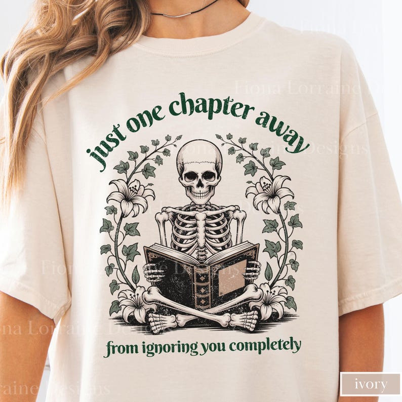 May include: Ivory-colored t-shirt featuring a skeleton reading a book, surrounded by floral and vine accents. The text reads "just one chapter away from ignoring you completely". The design is in shades of green and black.