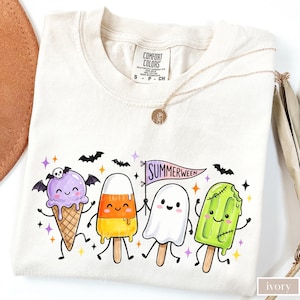 May include: An ivory t-shirt featuring a graphic of four cartoon treats: a purple ice cream cone with bat wings and a skull, a candy corn popsicle, a white ghost popsicle holding a Summerween flag, and a green Frankenstein popsicle.