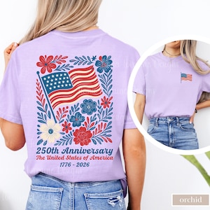 May include: A light purple t-shirt featuring a patriotic design. The back of the shirt displays an American flag with red and blue floral accents, and the text "250th Anniversary The United States of America 1776-2026". A smaller flag is on the front.