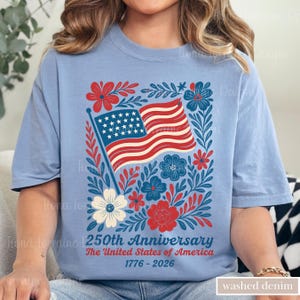 Puede incluir: Una camiseta azul claro con un dise&ntilde;o patri&oacute;tico. Presenta la bandera estadounidense, elementos florales rojos, blancos y azules, y el texto "250th Anniversary The United States of America 1776-2026". La camiseta est&aacute; etiquetada como "washed denim".