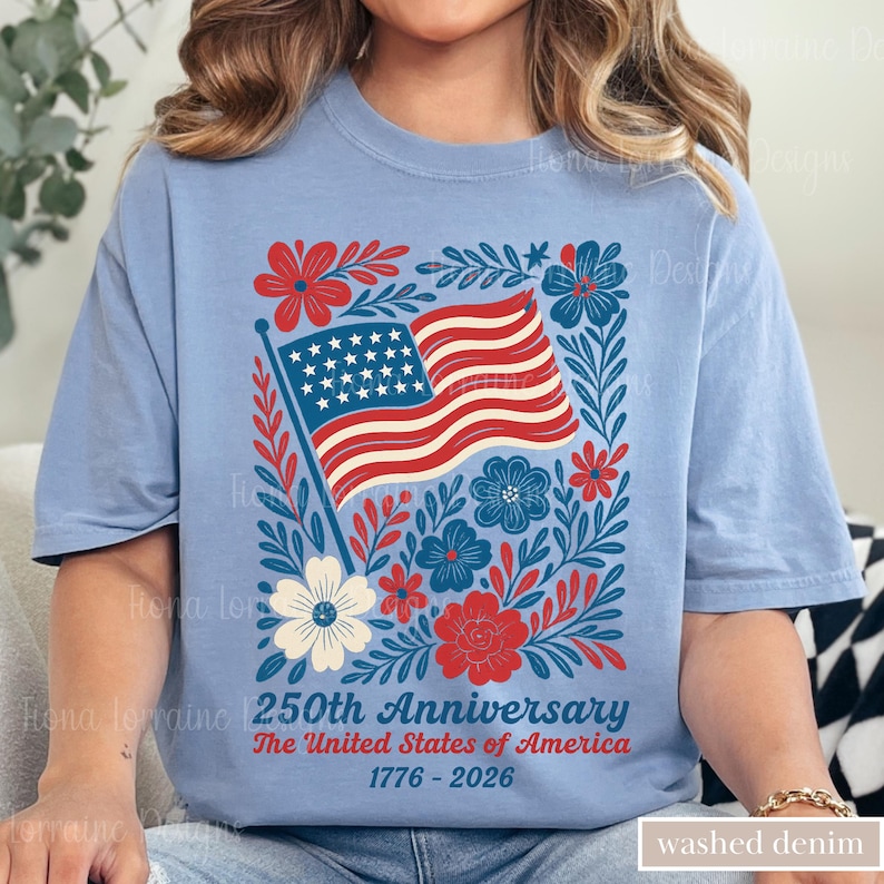 Puede incluir: Una camiseta azul claro con un dise&ntilde;o patri&oacute;tico. Presenta la bandera estadounidense, elementos florales rojos, blancos y azules, y el texto "250th Anniversary The United States of America 1776-2026". La camiseta est&aacute; etiquetada como "washed denim".