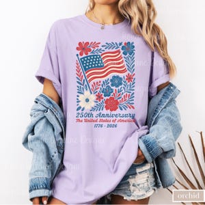 Puede incluir: Camiseta color orqu&iacute;dea con un dise&ntilde;o patri&oacute;tico para el 250 aniversario de los Estados Unidos. El gr&aacute;fico incluye una bandera americana, flores y texto. La camiseta es de un estilo c&oacute;modo e informal.