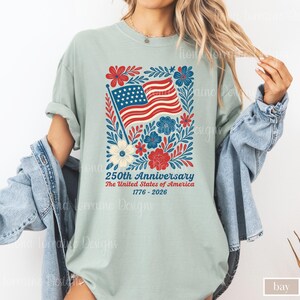 Puede incluir: Camiseta verde claro con un dise&ntilde;o patri&oacute;tico para el 250 aniversario de los Estados Unidos. El dise&ntilde;o incluye la bandera estadounidense, flores y el texto "250th Anniversary The United States of America 1776-2026."