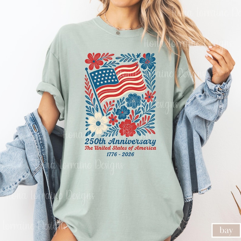 Puede incluir: Camiseta verde claro con un dise&ntilde;o patri&oacute;tico para el 250 aniversario de los Estados Unidos. El dise&ntilde;o incluye la bandera estadounidense, flores y el texto "250th Anniversary The United States of America 1776-2026."