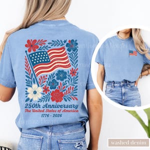 May include: Light blue t-shirt featuring a patriotic design for the 250th anniversary of the United States. The back of the shirt displays an American flag surrounded by floral elements. The text reads "250th Anniversary The United States of America 1776-2026".
