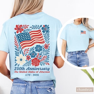 May include: Light blue t-shirt featuring a patriotic design. The back of the shirt displays an American flag with floral accents and the text "250th Anniversary The United States of America 1776-2026." A smaller flag is on the front.