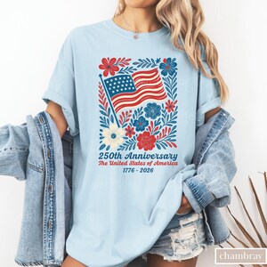 Puede incluir: Camiseta azul claro con un dise&ntilde;o patri&oacute;tico para el 250 aniversario de los Estados Unidos. El gr&aacute;fico incluye una bandera estadounidense ondeando, rodeada de elementos florales rojos, blancos y azules, con el texto "250th Anniversary The United States of America 1776-2026."