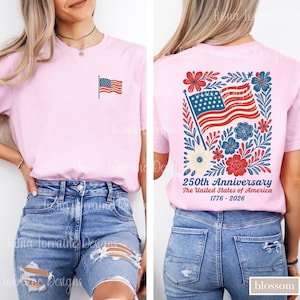 May include: Light pink t-shirt with a small American flag on the front and a larger design on the back. The back design features the American flag, flowers, and text that reads "250th Anniversary The United States of America 1776-2026."