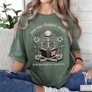 May include: Moss green t-shirt featuring a skeleton reading a book, surrounded by floral elements. The text reads "just one chapter away from ignoring you completely." The design is centered on the shirt.