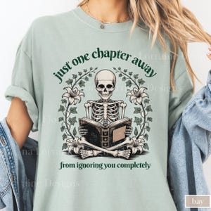 May include: A light green t-shirt featuring a skeleton reading a book, surrounded by floral elements. The text reads "just one chapter away from ignoring you completely". The shirt is a relaxed fit.