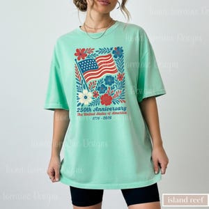 Puede incluir: Camiseta verde menta con un dise&ntilde;o floral que incorpora una bandera estadounidense y el texto "250th Anniversary The United States of America 1776-2026". El dise&ntilde;o incluye flores y hojas rojas, blancas y azules.