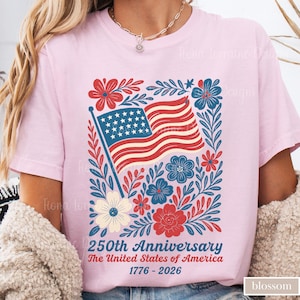 Puede incluir: Camiseta rosa claro con un dise&ntilde;o patri&oacute;tico. El gr&aacute;fico incluye una bandera estadounidense ondeando, rodeada de elementos florales rojos, blancos y azules. El texto dice "250th Anniversary The United States of America 1776-2026."