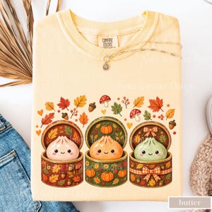 May include: A pale yellow t-shirt featuring a graphic of three round, smiling dumplings inside decorated containers. The design includes autumn leaves, acorns, mushrooms, and pumpkins in shades of orange, green, and brown. The shirt has a crew neckline.