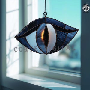 Puede incluir: Atrapasoles de vidrio manchado en forma de ojo, con tonos azul oscuro y blanco. El ojo tiene un contorno negro y está suspendido por una cadena negra. El texto "Cortes Digital" es visible.