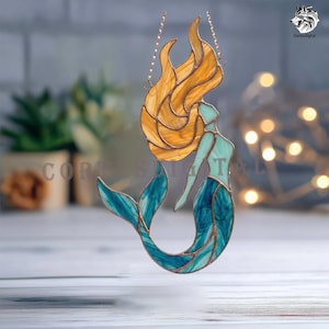 May include: A stained glass suncatcher shaped like a mermaid with flowing amber hair, a light mint green torso, and a deep blue tail. The figure is outlined in copper-colored metal solder and hangs from a delicate metal chain for display.