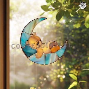 May include: Stained glass suncatcher depicting a sleeping teddy bear on a crescent moon. The moon is a mosaic of blue and teal glass, with a gold-coloured bear. Designed to hang in a window.