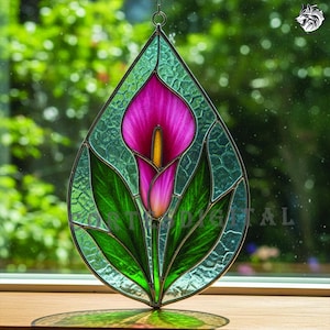 Op de afbeelding: Gebrandschilderd glaskunstwerk met een calla-lelie ontwerp. De lelie heeft een helder roze bloem met een gele meeldraad, omgeven door groene bladeren. Het glas heeft een druppelvorm met een getextureerde, lichtgroene achtergrond. Opgehangen aan een metalen ketting.