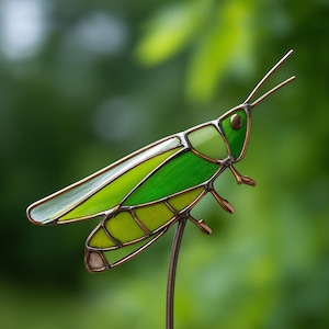 Puede incluir: Escultura de saltamontes de vidrieras con tonos verdes y acentos color cobre. El insecto tiene alas y patas detalladas, montado sobre una fina varilla de metal. El fondo es verde borroso.