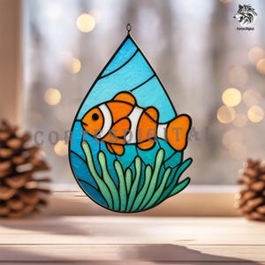 May include: Stained glass art piece featuring a clownfish in a teardrop shape. The fish is orange with white stripes, set against a blue background with green seaweed. The piece is designed to hang and is suitable for home decor.