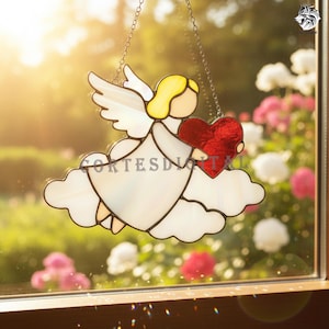 May include: Stained glass angel holding a red heart, suspended by a chain. The angel has yellow hair, white wings, and a white dress. The angel is set against a blurred background of flowers and sunlight.