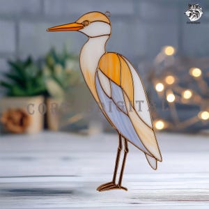 May include: Stained glass heron sculpture with a copper-colored frame. The bird features white, orange, and light blue glass panels. The heron has a long orange beak and stands on thin, brown metal legs. The text "CORTE DIGITAL" is visible.