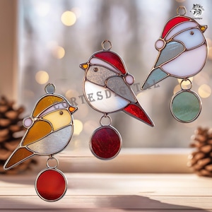 May include: Three stained glass bird ornaments, each wearing a red Santa hat. The birds have colourful wings and a round ornament hanging below. The background is blurred with soft lighting.