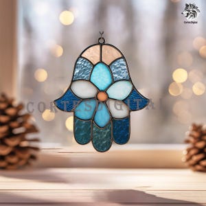 May include: Stained glass Hamsa hand suncatcher with a flower design in shades of blue, white, and orange. The suncatcher has a metal frame and is hanging in front of a window. The text "Cortes Digital" is visible.
