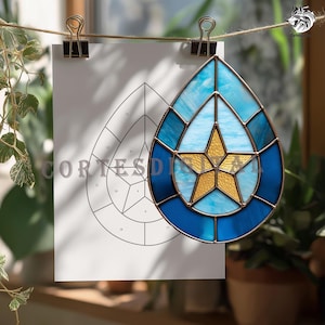 May include: A stained glass teardrop-shaped suncatcher with a gold star in the center, surrounded by shades of blue. The suncatcher is hanging with a paper pattern that says "CortesDia".