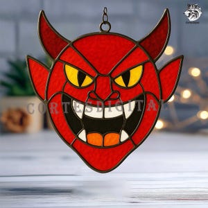 May include: A stained glass suncatcher shaped like a red devil face with yellow eyes, white teeth, and orange accents. The piece features dark metal lead lines and a small hanging loop at the top. The text CORTESDIGITAL is visible across the center.