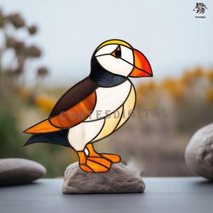 May include: A stained glass puffin bird sculpture with a colorful design. The bird features a white face, black and brown body, and orange beak and feet. It is perched on a small rock against a blurred outdoor background.