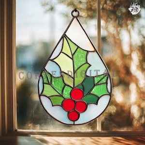 May include: Stained glass holly leaf and berry ornament. The teardrop-shaped decoration features green leaves, red berries, and a light blue background. The ornament is framed with a dark metal border and includes a hanging loop.