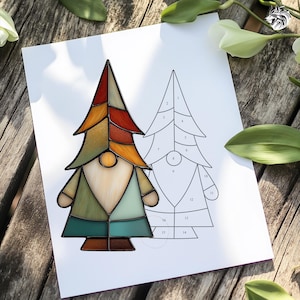 May include: A stained glass gnome in shades of green, orange, and brown, with a white beard and a round nose. A line drawing of the gnome is next to it. The artwork is on a white sheet of paper, resting on a wooden surface.
