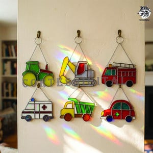 May include: Six stained glass suncatchers shaped like vehicles, including a green tractor, yellow excavator, red fire truck, white ambulance with a red cross, green dump truck, and red car, each hanging from a metal chain on a wall.