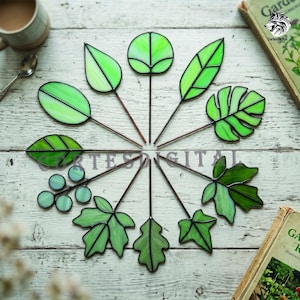 Puede incluir: Una colección de decoraciones de hojas de vidriera verde con tallos marrones dispuestas en un patrón circular. Las hojas varían en forma y tamaño, con contornos negros. La disposición está sobre una superficie de madera blanca. El texto "Cortes Digital" es visible.