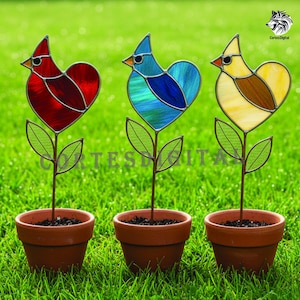 May include: Three stained glass bird garden stakes in terracotta pots. The birds are red, blue, and yellow with heart-shaped bodies and green leaves. The stakes are in a grassy yard.