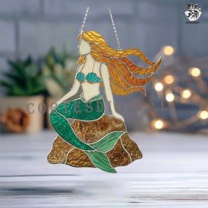 May include: A stained glass suncatcher depicting a mermaid with long flowing amber hair, a teal seashell top, and a textured green tail. The figure sits upon a brown rock formation, all outlined in dark metal solder, suspended by a silver chain.