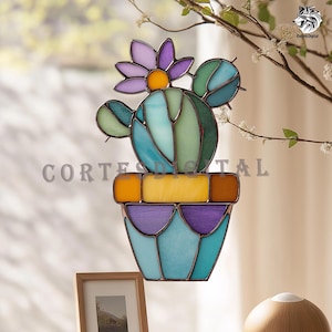 May include: Stained glass cactus window hanging with a purple flower, green and blue cactus, and a purple and blue pot. The text "Cortes Digital" is visible. The window art is displayed in front of a window.