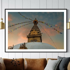 Könnte beinhalten: Eine weiße Stupa mit einer goldenen Spitze und Gebetsfahnen vor einem bewölkten Himmel bei Sonnenuntergang. Die Stupa hat ein großes Augenmotiv auf der Vorderseite.