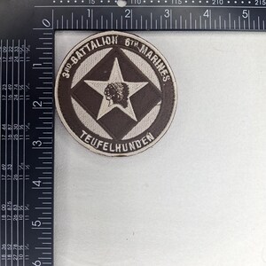 Pode incluir: Remendo circular com o texto "3rd BATTALION 6TH MARINES" e "TEUFELHUNDEN". O remendo apresenta uma estrela castanha e um perfil nativo americano sobre um fundo branco. O remendo tem aproximadamente 7,6 cm de diâmetro.