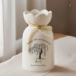 Puede incluir: Un jarrón de cerámica blanco con una parte superior festoneada y una cinta color crema atada alrededor del cuello. El jarrón tiene una ilustración negra de un sauce llorón y el texto "happy 9th anniversary", "A+E" y "Love Grows Here".