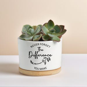Peut inclure: Un pot de fleurs en céramique blanche avec le texte "Never Forget The Difference You Make" en écriture noire. Le pot contient des plantes succulentes vertes et bordeaux et repose sur une base en bois ronde.