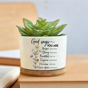 May include: A white ceramic planter containing a green succulent, featuring the text God says YOU ARE Unique, Strong, Beautiful, Forgiven, Precious, Never alone, accompanied by colorful floral illustrations and scripture references on the front.
