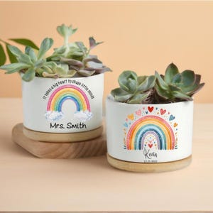 Könnte beinhalten: Zwei weiße Keramik-Pflanztöpfe mit Sukkulenten, jeweils mit Regenbogen-Design. Ein Topf trägt die Aufschrift "Mrs. Smith" und der andere "Kevin 12.25.2022". Beide Töpfe stehen auf Holzuntersetzern.